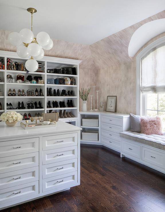 Luxurious white and pink walk-in closet is lit by a 10-light globe white glass and brass chandelier hung over a white island finished with drawers with polished nickel pulls. Behind the island, tall white shoe shelves are mounted against a wall clad in pink and gray wallpaper beside white corner shelves fixed next to a white built-in dresser. A storage window-seat is topped with a gray tufted cushion and pink sheepskin and gray pillows.