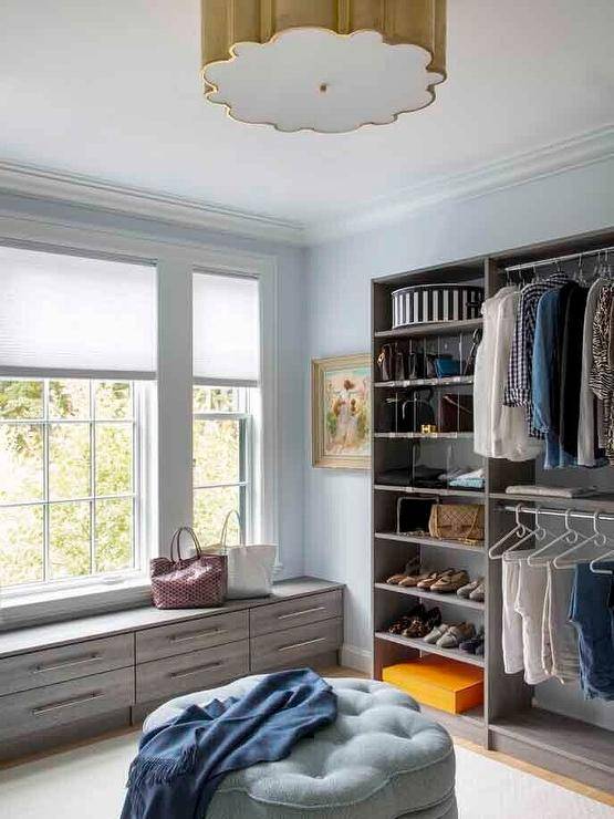 A blue tufted ottoman sits in a gray and blue custom walk-in closet lit by an Alexa Hampton Markos Grande Flush Mount. Stacked gray bench drawers are fixed beneath windows covered in white shades. Gray shoe and bag shelves fitted with acrylic dividers are built-in beside stacked clothing rails.