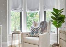 Contemporary nursery with a bay window furnished with an ivory wingback rocker and a fiddle leaf fig plant. Gray roman shades adorn the bay windows surrounded by white and gray wallpaper walls.