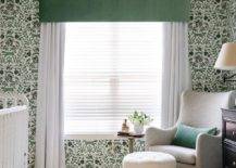 Green and white boy's nursery features a gray wingback glider with a white tufted pouf, a pedestal accent table, a cream rug, a green valance box over white curtains, green and white forest wallpaper and a white ceiling fan.