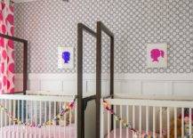 Contemporary nursery for fraternal twins boasts side-by-side white and brown cribs placed on a colorful geometric rug beneath silhouette art pieces hung from a wall clad in blue geometric wallpaper lined white board and batten trim.