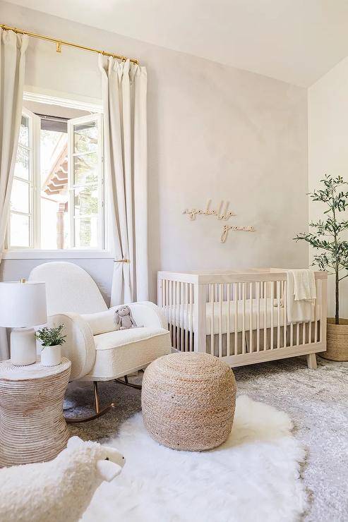 A soft pale pink boho chic nursery features gold letters on pale pink wallpaper over a light beige wooden nursery crib and a white boucle nursery rocker with tan ottoman and round accent table on white and gray layered rugs.