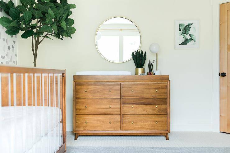 A round silver mirror hangs over a brown vintage dresser topped with a white changing pad. The dresser sits between a hung green abstract art piece and a fiddle leaf fig plant.