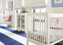 Spacious twin nursery design features a navy blue awning stripe rug, white wooden nursery cribs and a white glass front cabinet.