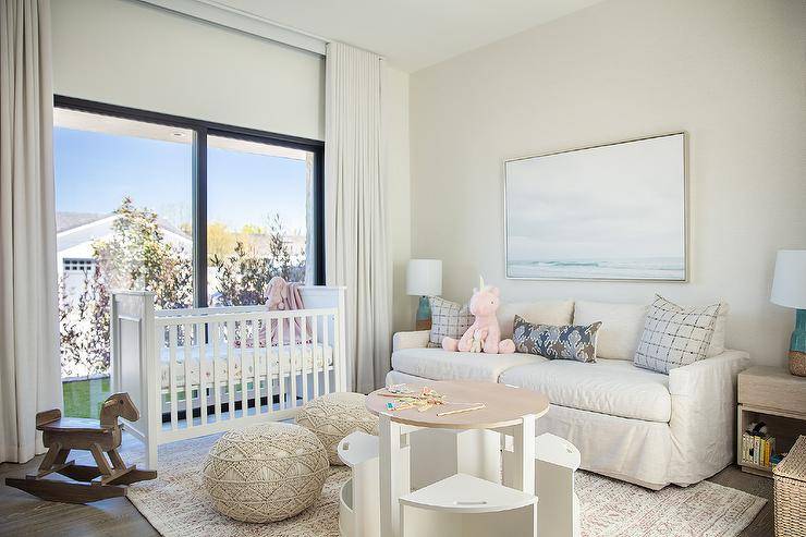 Welcoming nursery is styled with macrame poufs placed on a gray rug at a round two-tone play table with white chairs. n ocean art piece hangs over an off-white couch topped with gray and purple pillows lit by turquoise blue cork lamps. A white crib sits in front of sliding glass doors covered in white curtains.