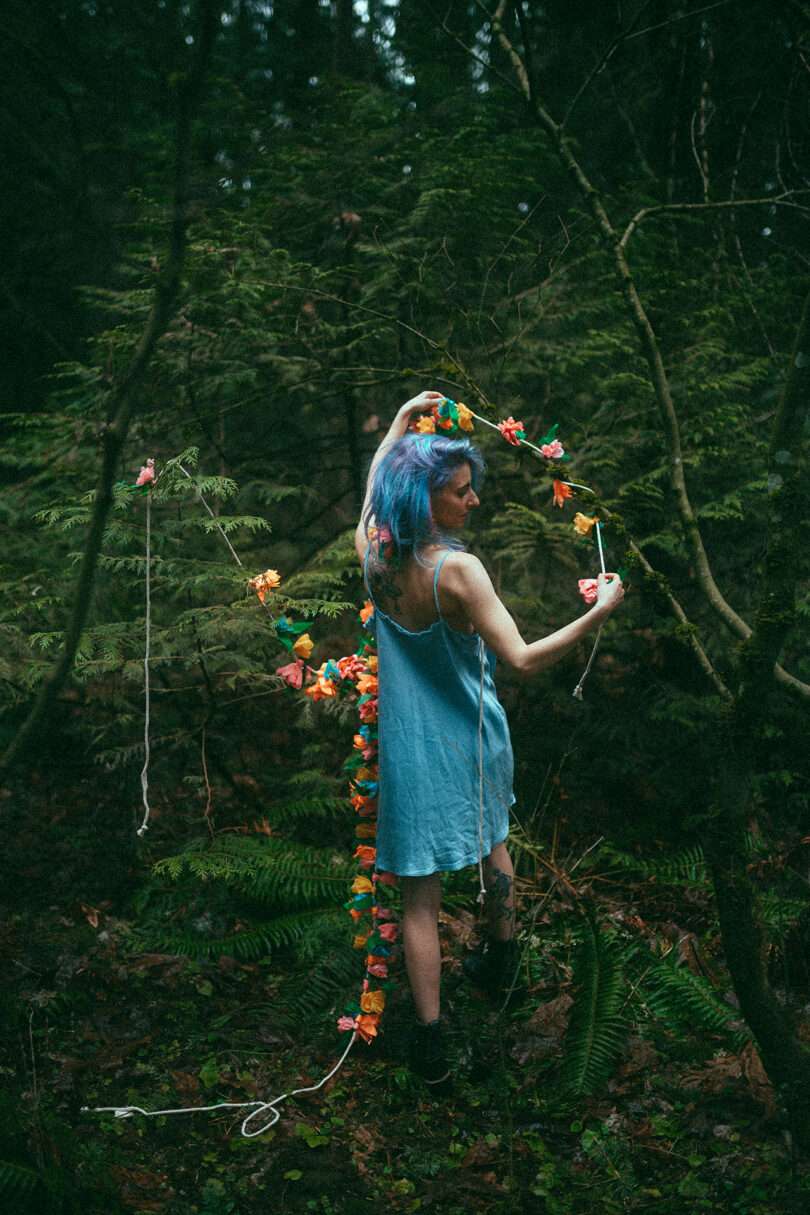 light-skinned woman with blue hair wearing a blue dress walks among greenery and florals