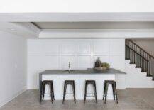 Four black Tolix stools sit in a basement at a white l-shaped island topped with a concrete waterfall countertop holding a round sink with a polished nickel faucet. The island is positioned in front of a wall covered in floor to ceiling wainscoting.