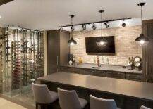 Contemporary basement bar features dark brown cabinets paired with gray quartz countertops and a brick backsplash lined with a flat panel tv illuminated by track lighting. Three industrial pendants hang over a dark brown bar island lined with gray bar stools placed next to a glass wine cellar.
