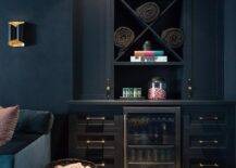 A black built-in basement bar features a glass front beverage fridge flanked by black cabinets donning black and gold pulls. The fridge is located beneath built-in x-shelves positioned between tall black shaker cabinets lit by a black 2-light sconce.