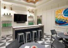Basement wet bar design features a gray waterfall bar with tolix stools on black and white geometric floor tiles, a tv flanked by floating shelves on board and batten trim and black chairs at a black round table.