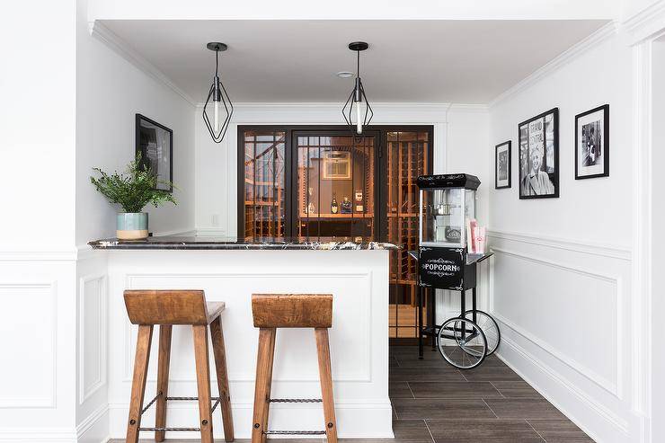 Basement wine cellar boasting a black marble countertop on a white peninsula finished with rustic wooden barstools and a set of sleek oil rubbed bronze pendant lights. Vintage popcorn cart adds charm to the corner of the wine cellar space.