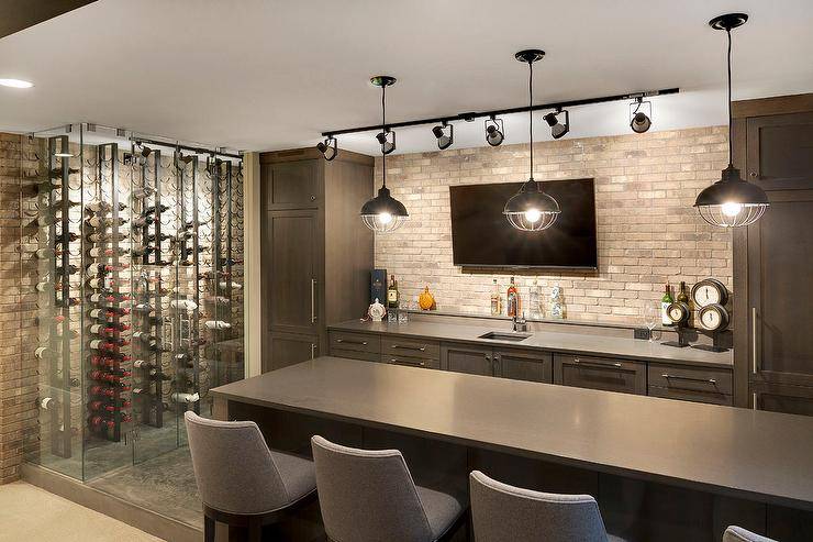 Contemporary basement bar features dark brown cabinets paired with gray quartz countertops and a brick backsplash lined with a flat panel tv illuminated by track lighting. Three industrial pendants hang over a dark brown bar island lined with gray bar stools placed next to a glass wine cellar.