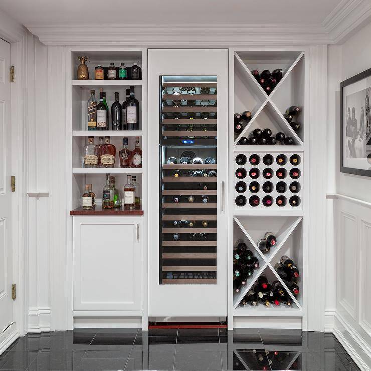 Basement with built-in wine racks and white cabinets finished with nickel hardware storing wine and beverages and spirits. This custom basement wine room features a tall wine fridge centered between shelves.