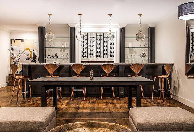 A black wet bar is topped with white marble lined with Cherner Barstools illuminated by four glass globe pendants. Chic basement wet bar boasts a row of vertical wine racks flanked by glass shelves.