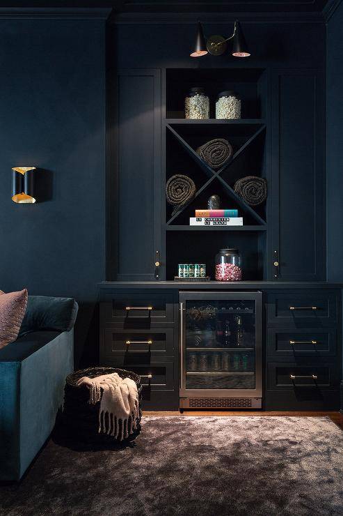 A black built-in basement bar features a glass front beverage fridge flanked by black cabinets donning black and gold pulls. The fridge is located beneath built-in x-shelves positioned between tall black shaker cabinets lit by a black 2-light sconce.