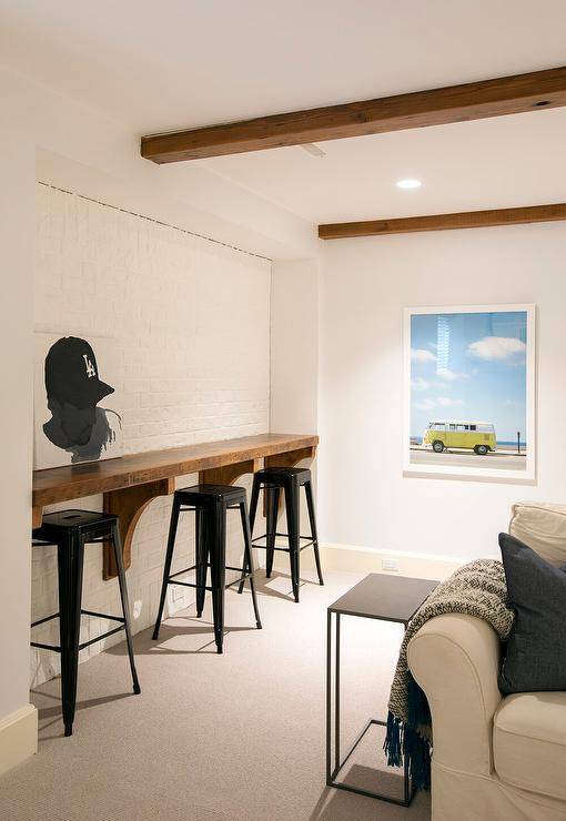 Basement family room is fitted with stained wood ceiling beams mounted over a stained wood built-in bar fixed against a white brick wall and matched with black Tolix stools.