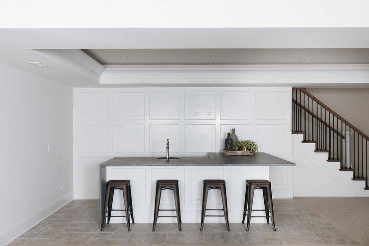 Four black Tolix stools sit in a basement at a white l-shaped island topped with a concrete waterfall countertop holding a round sink with a polished nickel faucet. The island is positioned in front of a wall covered in floor to ceiling wainscoting.