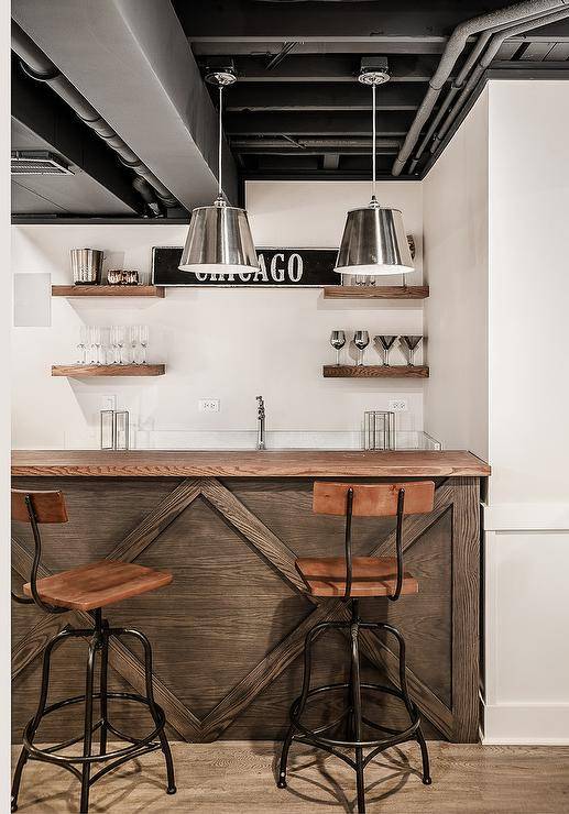 An industrial style basement features a distressed coffee stained oak wood bar counter with metal and wood swivel stools under silver metal light pendants. Stained oak floating wet bar shelves flank "Chicago" sign under exposed pipe ceilings.