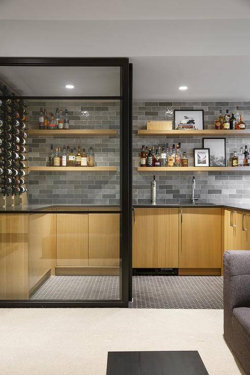 Sliding glass doors open to a basement wet bar featuring beige veneer cabinets complemented with a gray quartz countertop and a sink with a polished nickel gooseneck faucet. The faucet is mounted beneath beige floating shelves fixed against gray offset tiles lit by recessed lighting.