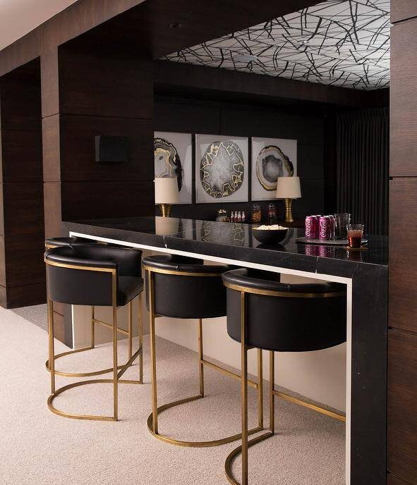 Three stunning Arteriors Calvin Barstools sit on a carpeted floor in front of a black marble waterfall bar in this well appointed contemporary basement.