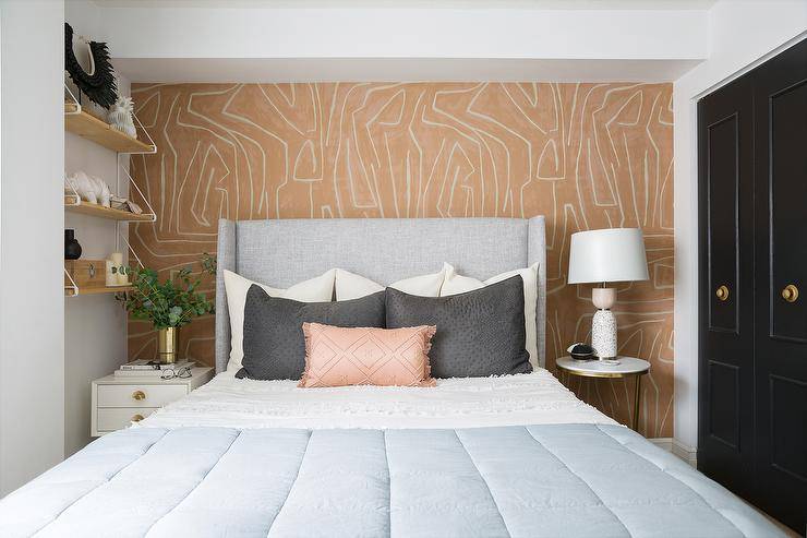 Contemporary bedroom features a dove gray wingback bed on orange Groundworks Graffito wallpaper and mismatched bedside tables.