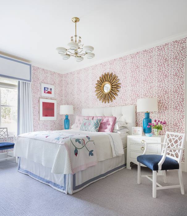 Contemporary bedroom features a white tufted headboard accented with pink pillows flanked by white nightstands lit by Caribbean blue lamps, a white chair with blue cushion and a gold sunburst mirror mounted on Brunschwig & Fils les touches pink wallpaper.
