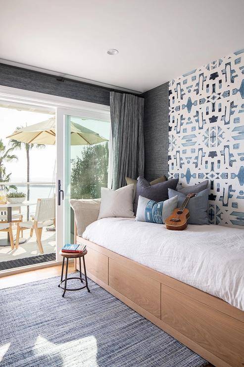 Charcoal gray curtains cover sliding glass doors surrounded by charcoal gray wallpaper in a bedroom featuring a custom built daybed fitted with drawers and mounted against an accent wall clad in blue trellis wallpaper. The bed sits on a blue jute rug.