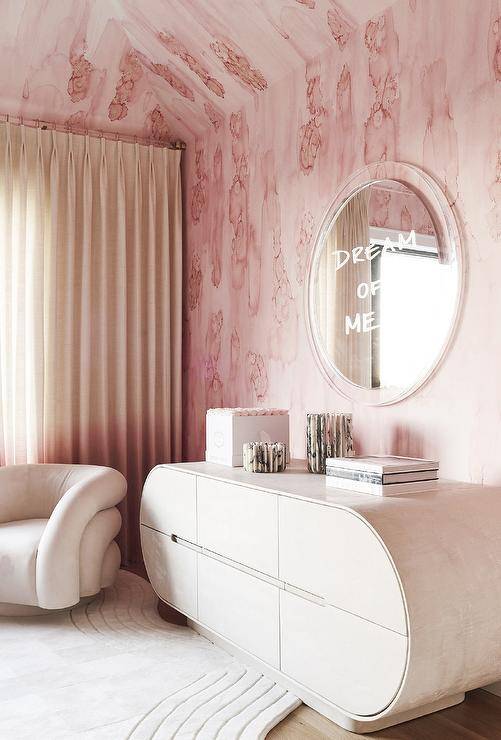 Charming contemporary bedroom features walls covered in pink wallpaper and a "Dream of Me" mirror hung above an oblong credenza, as a light pink velvet accent chair sits in a corner on a cream rug.