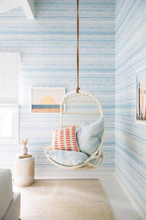 Charming white and blue bedroom features a white rattan hanging chair topped with a blue chevron and orange pillow and hung in a corner in front of walls coered in blue striped wallpaper.