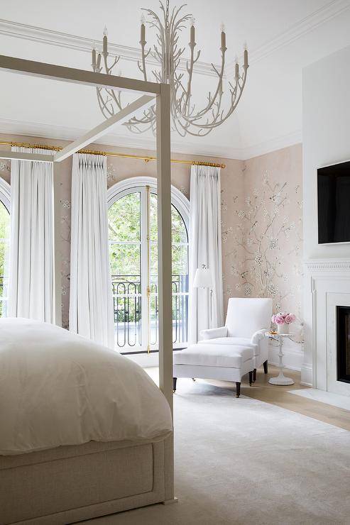 A white faux bois chandelier lights a bedroom clad in pink chinoiserie wallpaper and boasting a light gray canopy bed placed on a light gray rug. Tall arched windows are covered in white curtains hung from gold rods, while a white corner chair and ottoman sit in a corner beside a white spindle accent table.