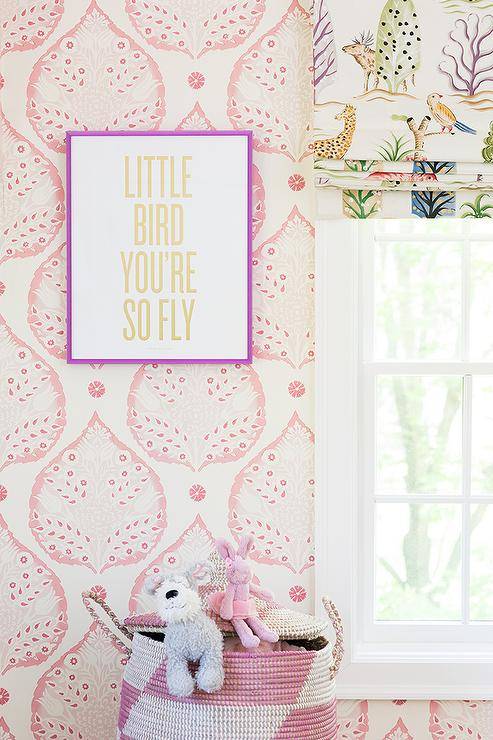 A yellow typography print in a purple frame hangs in a gorgeous girls' bedroom from a wall clad in Galbraith & Paul Lotus Wallpaper over a white and pink Moroccan basket. A window is covered in an animal print roman shade.