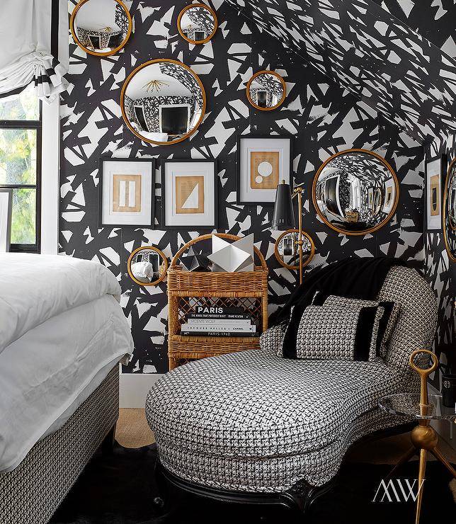 Chic contemporary bedroom is covered in black strokes print wallpaper and features a stunning black and white houndstooth chaise lounge placed on a black cowhide rug. Tan and white abstract art is hung in black frames is hung beneath and beside gold framed round convex mirrors.
