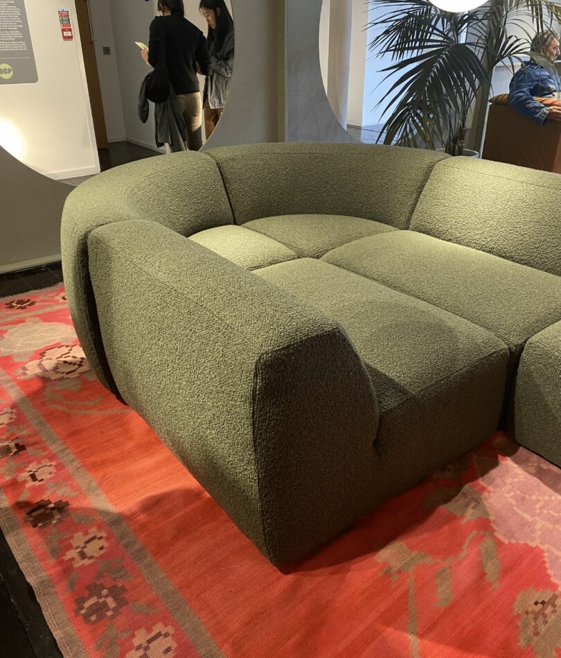A series of sofa modules in olive green combine to create a semicircular enclosed seating put. A red rug is beneath and some people can be seen in the background.