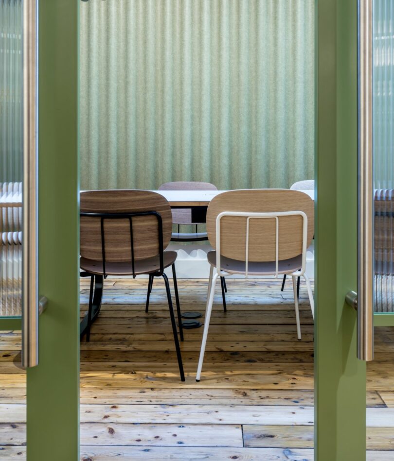 A meeting room with table and chairs can be seen through two green-framed reeded glass doors. The back wall is covered with a corrugated acoustic panel. 
