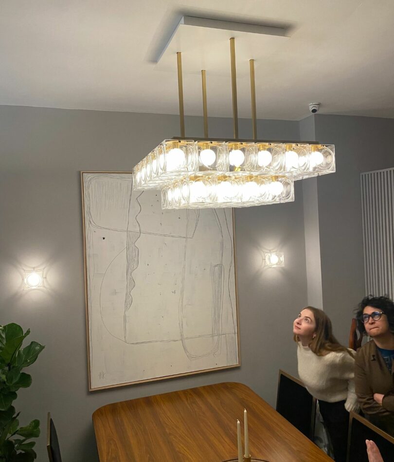 A white woman in her 20s and an Indian woman in her 40s lean over a table and up to the underside of a flat, square chandelier made of a layer of lightbulbs encased within tessellating square blocks of glass.