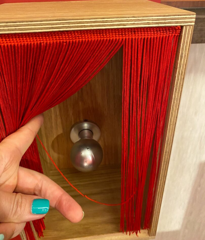 A white finger draws aside a red string curtain on the front of a wooden box to reveal a doorknob. 
