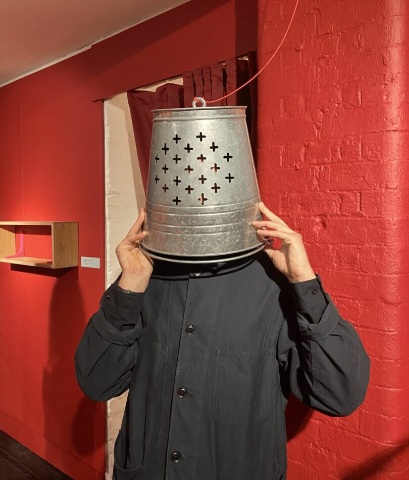 A white man in a black shirt stands in front of a red wall with an upturned bucket on his head. The bucket has a series of perforations that mimic the holes in a confessional booth. 