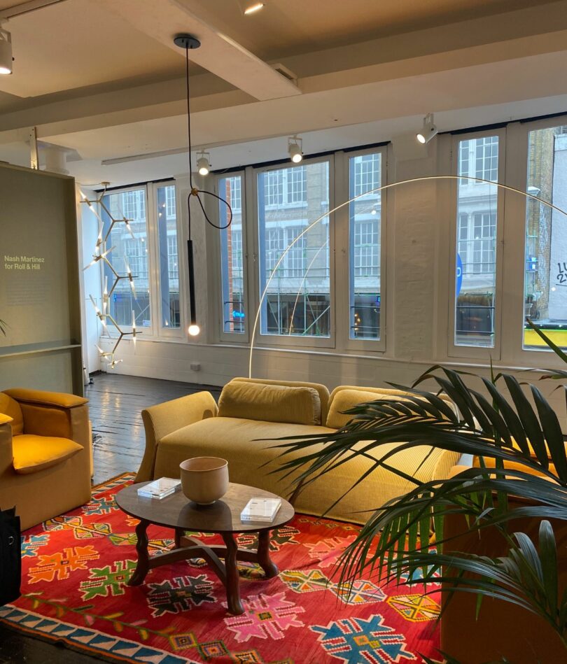 A showroom includes a yellow sofa and armchair, red patterned rug, a plant in the foreground, a coffee table and pendant light. Windows in the background look out on a building beyond. 