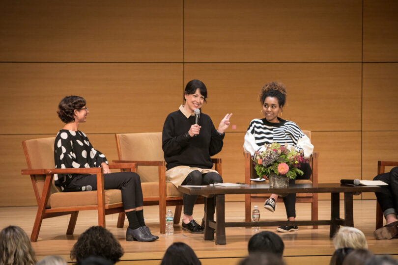 three light-skinned people with dark hair sitting in chairs on a stage having a discussion
