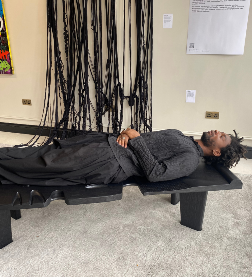 Giles Nartey, a black man in his 30s, lies on a black wooden daybed in front of a braided, knotted and tied wall handing. 