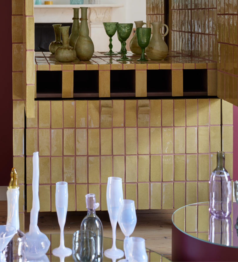 A cabinet made from yellow tiles with pink grout is open. Inside is a mirror and green glasses and vessels. On mirrored tables in front are white and lilac glasses and bottles. 