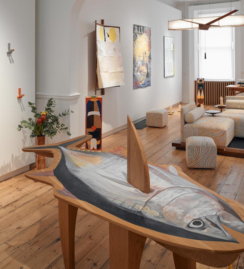 A table shaped like a fish with a fin piercing the surface is in the foreground of a large room populated with other pieces of art and furniture. 