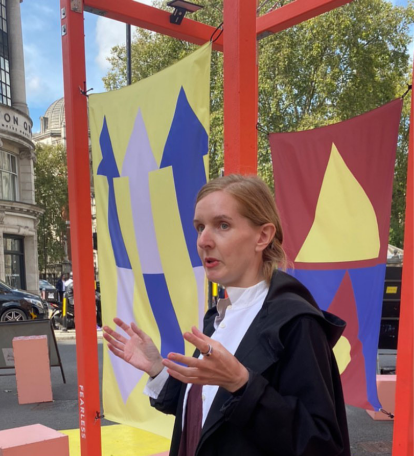 A frame of red-painted wooden beams supports a series of graphic flags in front of which stands Eva Feldkamp, a white women in her 40s, captured mid-word. 