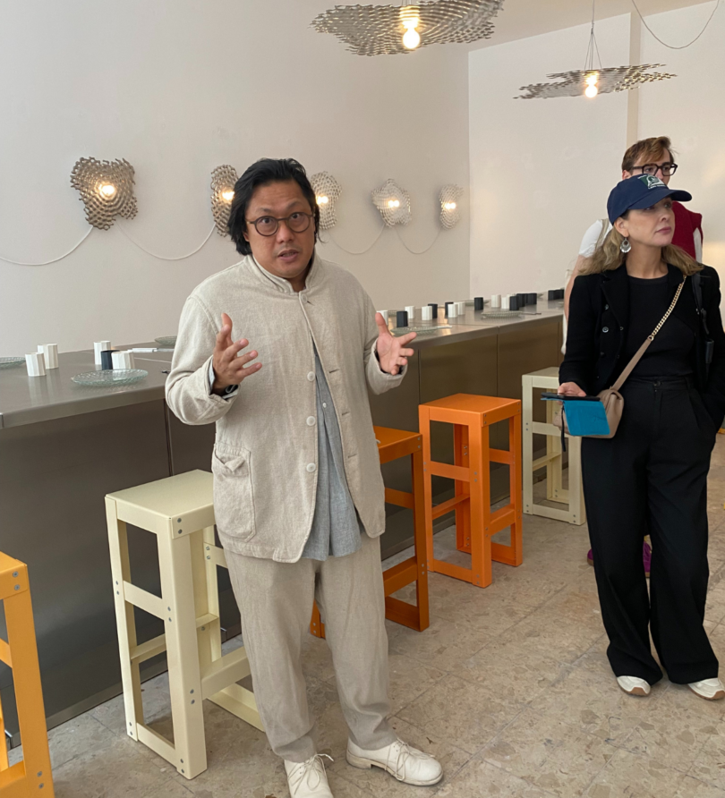 Guan Lee, an Asian man in his 50s, wears a pale linen suit and stands in front of a long table and a series of multi-coloured high stools. He is captured mid-sentence, gesticulating with his hands. 