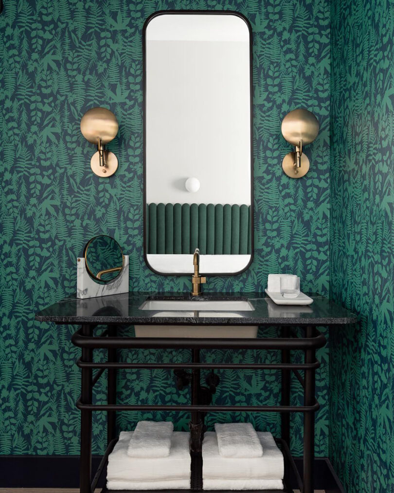 Bathroom with green and black wallpaper, long vertical mirror with brass circular sconces on each side, and black marble pedestal sink. A pile of folded white bath towels are underneath the sink on a small shelf.