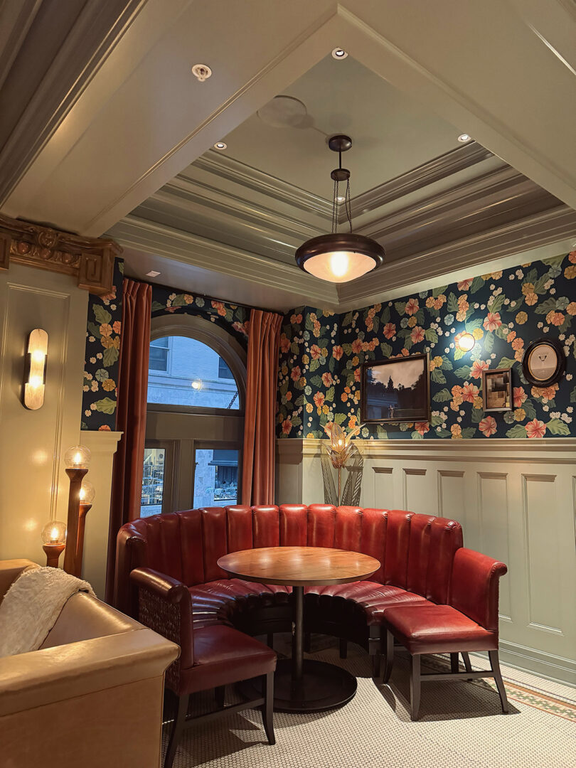 Corner half-circle dining booth inside hotel's Abigail Hall, with historic-styled pendant glass lighting overhead circular table and floral wallpaper over wainscoting detail.