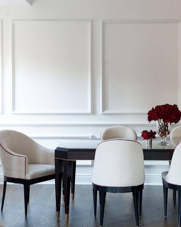 White French fabric dining chairs surround a glossy black French dining table placed in front of wainscot walls.