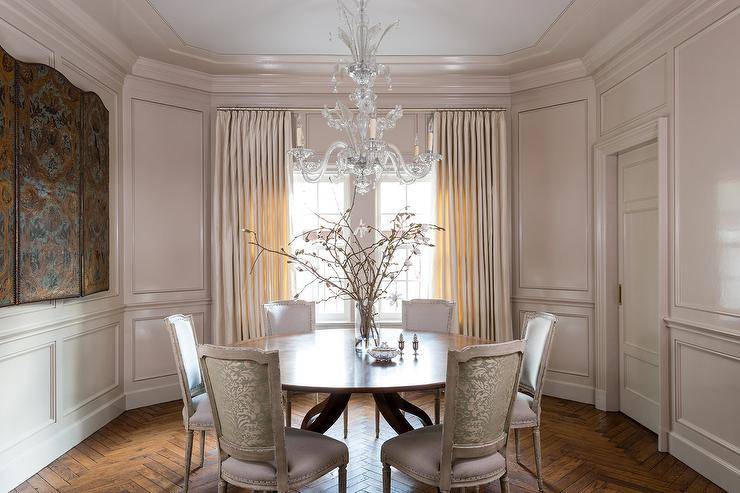 Elegant French dining space boasts a crystal chandelier fixed over a round dining table surrounded by pink and green damask French dining chairs positioned on a herringbone wood floor. Pale beige wainscot walls are lined with matching crown molding, while windows are covered in light beige curtains.