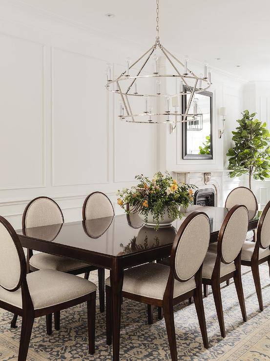 In front of a wainscot wall, gray and brown oval back dining chairs are placed on an ivory and blue wool rug around a glossy brown lacquer dining table lit by a 2-tier nickel candelabra chandelier.
