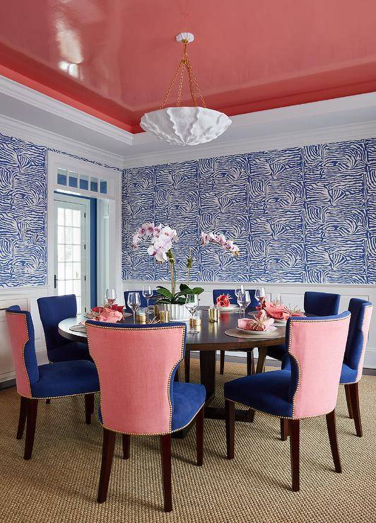 A glossy pink gray ceiling complements a round brown wooden dining table paired with blue and pink dining chairs placed on a natural woven rug. Wainscot walls are finished with Brunschwig & Fils Ashanti Wallpaper.